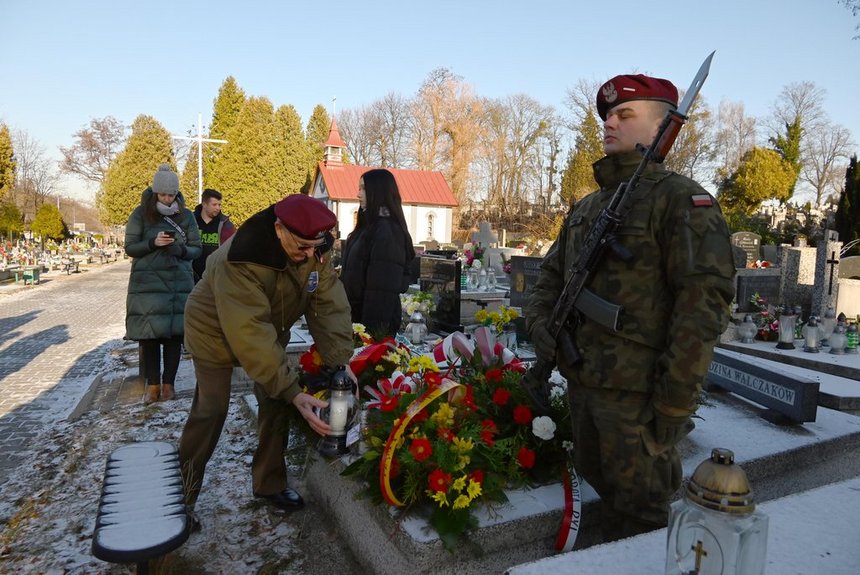 Cmentarz. Przy grobie weteranów powstania Wielkopolskiego stoi warta honorowa. Na pierwszym planie żołnierz na baczność z karabinem i bagnetem. Na grobie kwiaty. Drugi żołnierz stawia na grobie zapalony znicz. W tle widzać tablicę grobową na napisem Rodzina Walczaków.