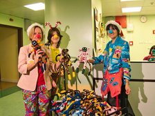 Na zdjęciu widać trzy dorosłe osoby przebrane w kolorowe stroje klownów. Stoją w szpitalnym korytarzu obok wózka pełnego pluszowych, materiałowych małpek.. Mają czerwone nosy i czapki, uśmiechają się i trzymają materiałowe maskotki w rękach