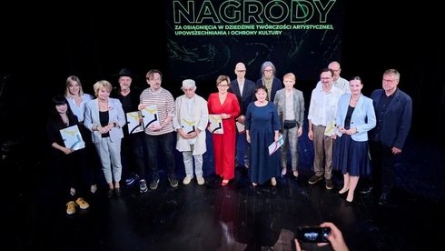 Na scenie teatralnej stoi grupa ludzi, którzy w rękach trzymają dyplomy. Są to laureaci nagród w 2023 roku. W tle napis: Nagrody za osiągnięcia w dziedzinie twórczości artystycznej, upowszechnia i ochrony kultury.