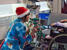 Na zdjęciu widzimy dorosłą osobę w czapce Mikołaja, kucającą przy dziecku na wózku inwalidzkim, w rękach trzyma i pokazuje dziecku dwie kolorowe materiałowe maskotki w kształcie małpek. W tle widać sprzęt medyczny i monitor.