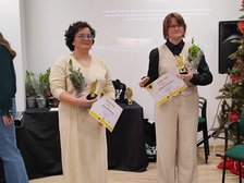 Na zdjęciu widzimy dwie osoby w jasnych strojach trzymajace w dłoniach statuetki, kwiaty oraz dyplomy. Zdjęcie zrobione w pomieszczeniu na tle białego ekranu.