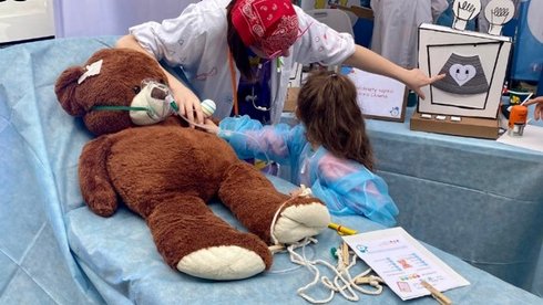 Na zdjęciu widać stoisko Fundacji Dr Clown, organizacji znanej z działań na rzecz uśmiechu i wsparcia emocjonalnego dzieci w szpitalach. Na pierwszym planie znajduje się duży pluszowy miś leżący na niebieskim łóżku szpitalnym – wygląda, jakby był „pacjentem”. Obok niego stoi dziewczynka w niebieskim fartuchu ochronnym, która „bada” misia razem z dorosłą osobą w białym fartuchu, z czerwoną chustką na głowie i kolorowymi elementami stroju. W tle widać banery z logo Fundacji Dr Clown i hasłem „#uśmiechleczy