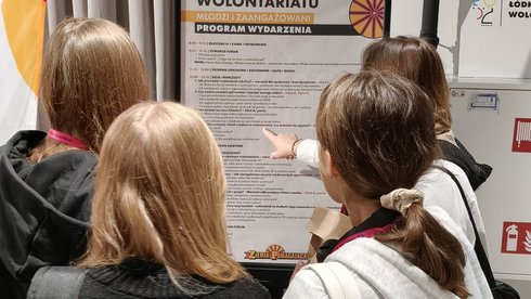 Na zdjęciu widzimy grupę młodzieży wskazującą plakat, który znajduje się przed nimi na staludze. Na plakacie wielkimi literami napisane jest: Forum Łódka Wolontariatu. Młodzi i Zaangarzowani. Program Wydarzenia. 
