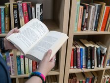Dłonie trzymające otwartą książkę na tle regału bibliotecznego.