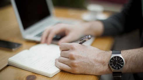 Zbliżenie na męskie dłonie trzymające długopis. Na lewej ręce czarny zegarek. Mężczyzna siedzi przy biurku i notuje coś w brulionie, w tle srebrny laptop z wyłączonym ekranem.